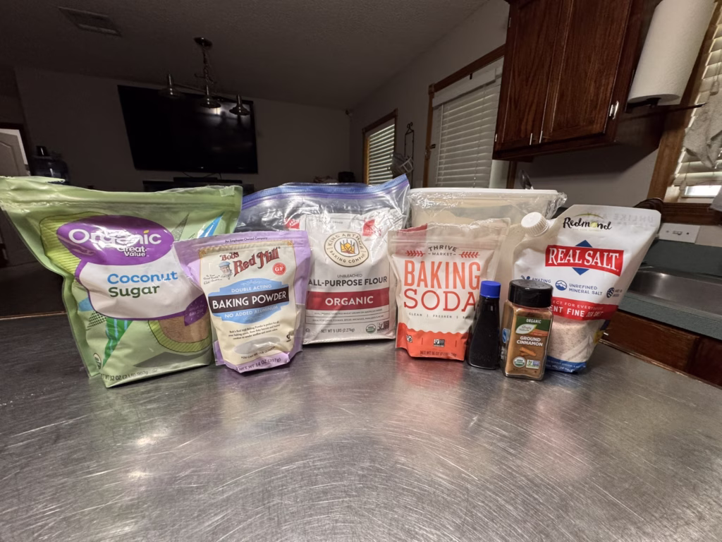 A lineup of clean baking staples including coconut sugar, flour, baking powder, baking soda, cinnamon, vanilla, and mineral salt on a kitchen counter