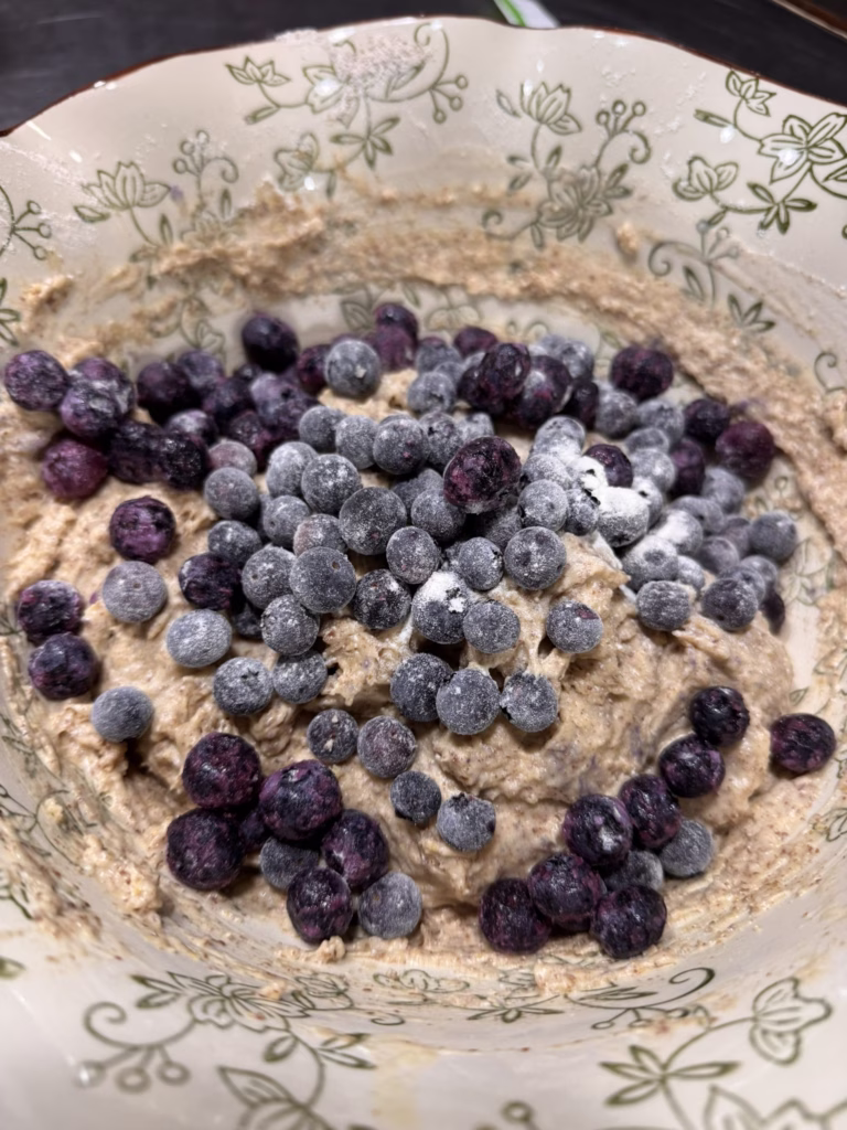 Frozen blueberries tossed in flour sitting atop whole-food muffin batter in a floral mixing bowl