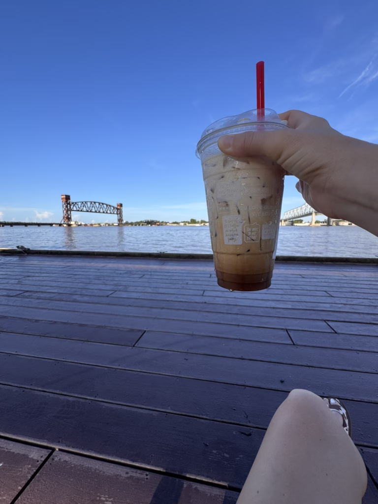 A hand holds a large iced coffee cup with a red straw, overlooking a calm body of water with a distant bridge under a bright blue sky.