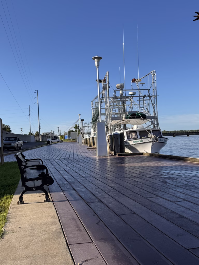 Shrimp boats are docked along a wooden riverfront promenade with a black bench in the foreground under a clear blue sky.