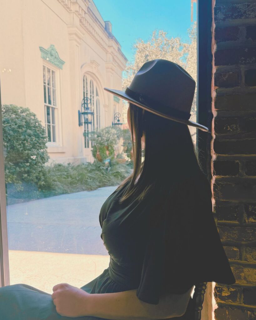 Woman in a wide-brimmed hat sits in quiet reflection by a sunlit window, looking out toward a garden courtyard; the scene feels peaceful, grounded, and introspective.